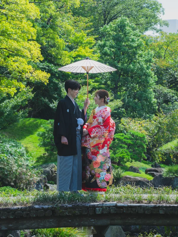 庭園の池にかかる橋の上で、白い番傘を持つ赤い色打掛の花嫁と新郎