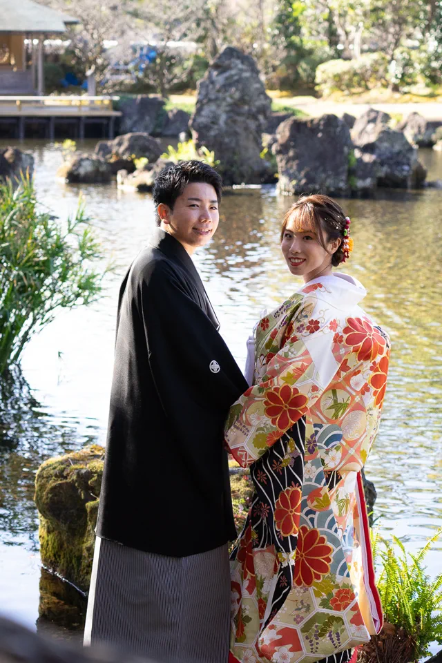 庭園の池と石組みを背に寄り添う、色打掛の花嫁と新郎