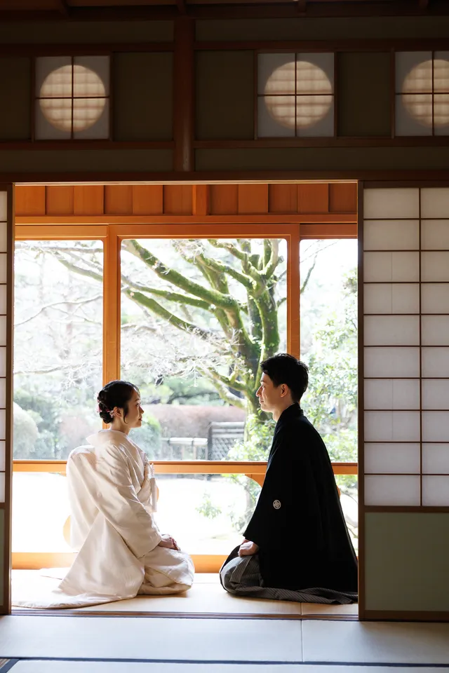 丸窓のある茶室の座敷で、開いた障子の先の庭に向かって座る白無垢の花嫁と新郎