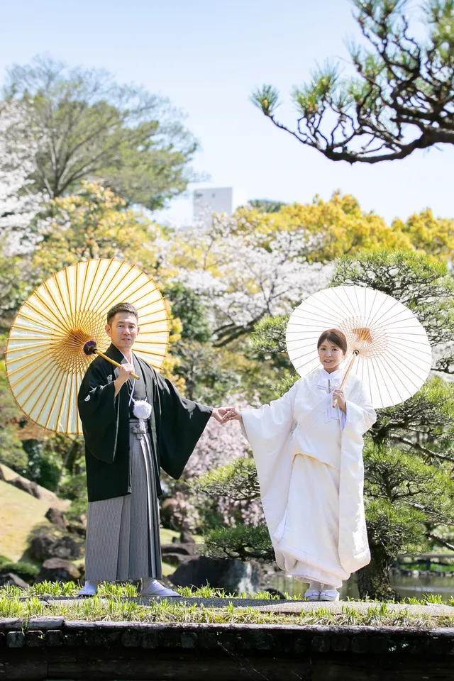 松のある庭園で、番傘を持つ白無垢の花嫁と新郎