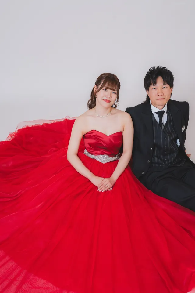 灰色の背景のスタジオで、赤いカラードレスを広げて座る花嫁と新郎