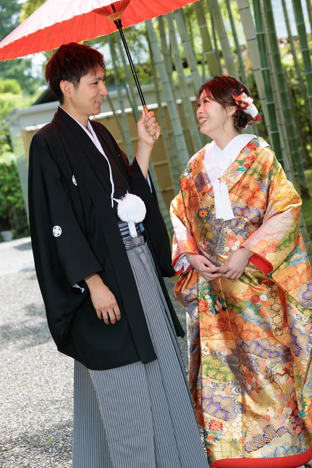竹林で赤い番傘をさす、朱と金の色打掛の花嫁と新郎