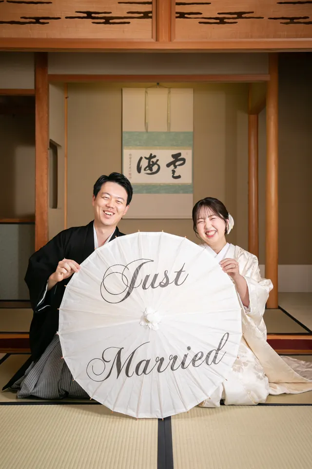 掛け軸の座敷で、Just Married と書いた番傘を持って座る白無垢の花嫁と新郎